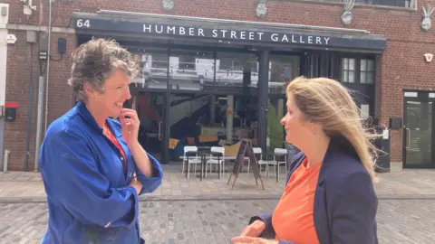 BBC / Sally Fairfax Lady smiling with short wavy hair, blue denim jacket and striped red and orange jumper with lady with long brown hair, blue jacket and orange top.
They are standing on a paved street in front of a brick and glass building with tables and chairs outside of it and an advertising board outside.