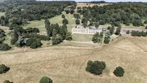 A drone view of Ashton Court estate with large areas of grassland showing light brown instead of green. Small trees are dotted around the landscape and the lines of footpaths through the landscape are also visible