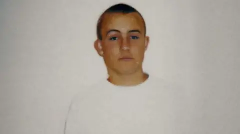 Police Scotland/ Firecrest Films Police photograph of a teenage boy looking blanky at the camera against a white wall. He has short blond cropped hair and is wearing a long-sleeved white top.