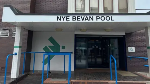 Nye Bevan swimming bath building. It is a dark brown brick building. The front is made up of green and white tiles and automatic glass doors. There are blue railings and also a ramp alongside steps leading up to the entrance.