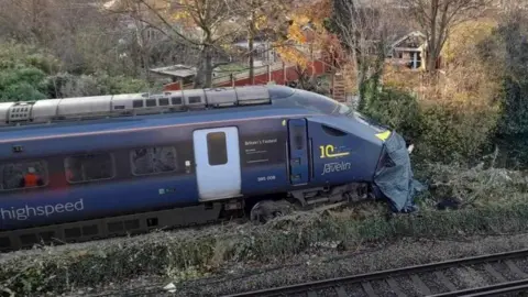 A blue train with the words high speed written on the side in grey. The train has left the track and is in green undergrowth. Trees are in the background