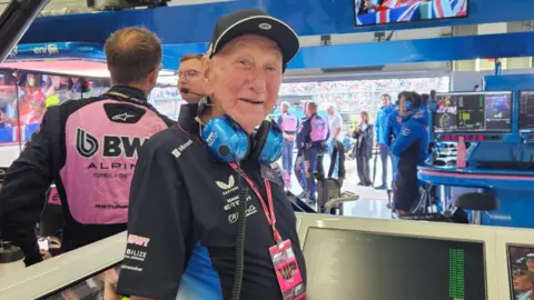 An older man wearing a black cap, blue headphones, and a black shirt stands in a motorsport pit area. Behind him, team members in pink and black uniforms work near monitors, with spectators visible through the open garage.
