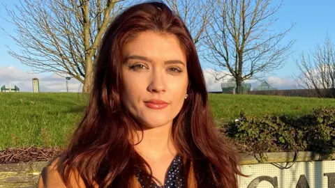 BBC Cara Hunter standing in front of a grass will with trees on top. 