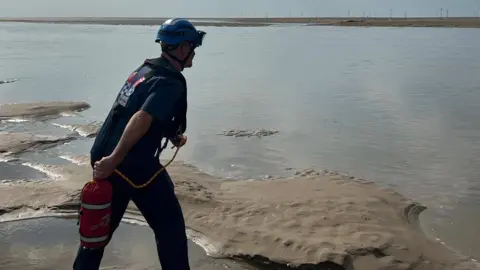 A rescue officer in blue uniform and helmet appears to throw out a safety line from the sands acorss the water at Leasowe.