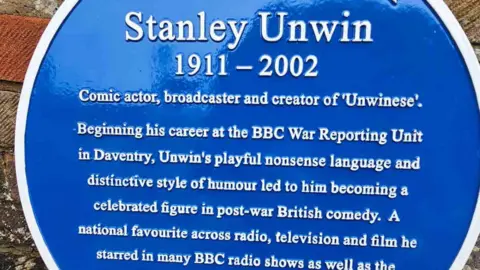Kate Bradbrook/BBC The blue plaque unveiled at Stanley Unwin's old house. It reads: "Stanley Unwin. 1911-2002. Comic actor, broadcaster and creator of Unwinese. Beginning his career at the BBC War Reporting Unit in Daventry, Unwin's playful nonsense language and distinctive style of humour led to him becoming a celebrated figure in post-war British comedy. A national favourite across radio, television and film he starred in many BBC radio shows." The rest of the words are cut out of the picture.