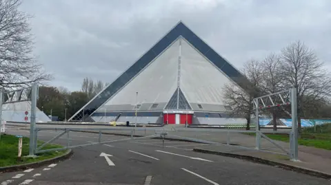 A triangle shaped building surrounded by a deserted car park and a closed gate at the entrance.