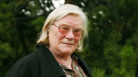 Getty/Ulfanderson Shows Jennifer Johnston wearing glasses in a black coat and standing in front of trees