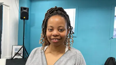 A woman with curly, dreadlocked hair wearing a grey tracksuit top.