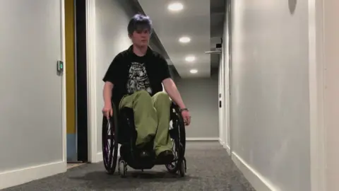 BBC/Seb Cheer A person wearing green trousers and a black top sits in a wheelchair and wheels themself out of a door, heading down a corridor to another door.