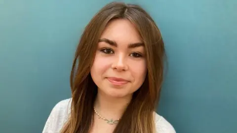 Alex Pope/BBC Amie, standing in front of a green wall, looking straight at the camera. She has long dark hair, with a middle parting, and has on a necklace and a grey top.
