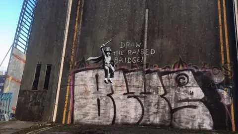 A painted mural depicting a small boy is sitting on top of large letters wearing a cape and holding a wooden sword with a pencil stuck to the top of it. A caption next to the image reads "Draw the raised bridge!"