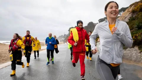 RNLI / Nathan Williams People running near the sea edge - a woman at the front is wearing a RNLI Lifeboats top