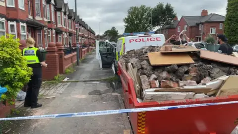 Skip filled with bricks and rubble outside in street taped off by police