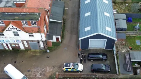 An aerial view of the site in Eastbourne shows a police car near the units and a group of officers nearby. There are different types of buildings on the site, some with signs on the front and red roofs and other newer buildings with garage doors.