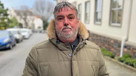 A man with black and grey hair loking at the camera. He is wearing a green jacket with a fluffy hood. 