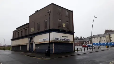A derelict looking building sits on the junction of a road with a carpark behind. The front is three floors and the rear two and it is built of brick which is painted a dark brown-grey. The lower section is rendered or clad in white. There are huge blue metal shutters pulled down over what would be large windows. White shop signs show - in large blue letters - it was once home to High Street Tyres. The backs of other buildings can be seen off to the right, but the tyre shop sits very much alone in the image.