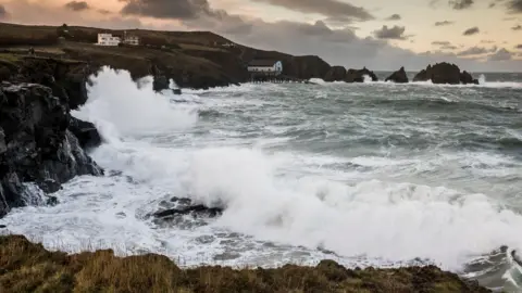 PA A side view of a coastline with crashing waves and a rocky peninsula of land with a few houses on.