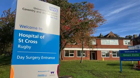 BBC A blue and silver NHS sign that says Welcome to Hospital of St Cross Rugby, in front of a red brick building and a tree with autumnal leaves