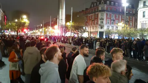 PA Crowds lining the streets in Dublin City Centre, with the Dublin Spire in the background. It is night time, and the streets are lit.