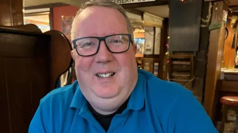 Image showing head and body shot of Stuart McMahon from the Campaign for Real Ale. He has short, light hair and is wearing black-rimmed rectangular glasses. He has on a bright blue polo shirt with a white CAMRA logo on the left hand-side chest area.