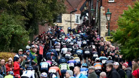 Getty Images/George Wood Riders at the Lincoln Grand Prix head up Michaelgate towards the cathedral and Castle Square. Large crowds are standing at each side of the cobbled road with the cyclists in the middle.
