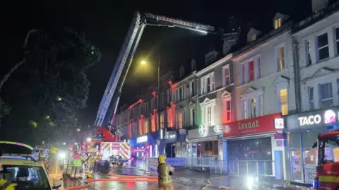 A firefighter watches on as a ladder crew extinguish the fire. The street is full of lit up bars and fast food stores.