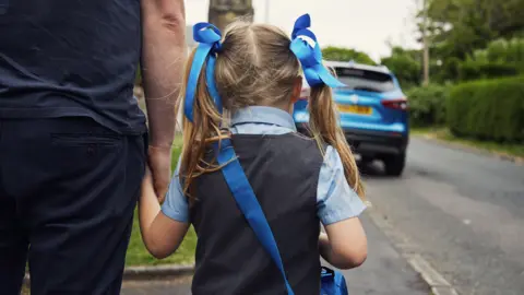 Getty Images The back of a small child wearing a grey dress over a pale blue school shirt. She has a deeper blue bag over her shoulder and blue ribbons in her long fair hair. The girl is walking along the road holding a man's hand. 