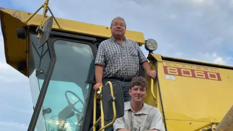 Jon Watt Jon Watt sitting on the steps of a yellow combine harvester. He has short mid-brown hair, curling on top, and is wearing a checked shirt. Behind him his father is standing in the doorway of the combine's cab. He has short grey hair and is wearing glasses, a grey checked shirt and grey trousers.