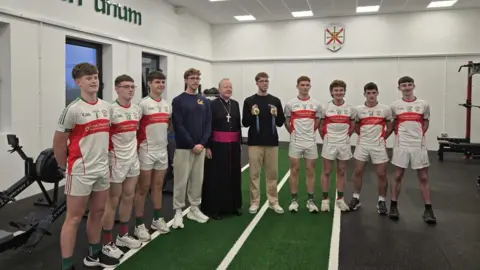 Daniel Wiffin School pupils in white and red sports kits stand beside Daniel and Nathan Wiffin. Daniel wears beige trousers and black top and holds his Olympic medals. Nathan wears grey trousers and a navy sweatshirt. The archbishop, wearing a black robe and a cross, stands between them.