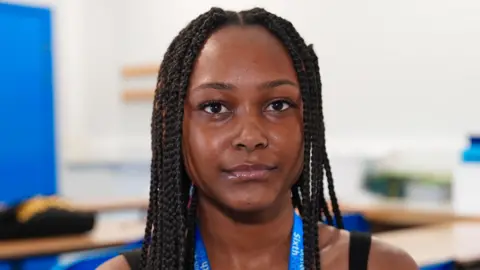 A 16-year-old girl with black braided hair is looking at the camera. She is wearing a black dress and has a blue school lanyard
