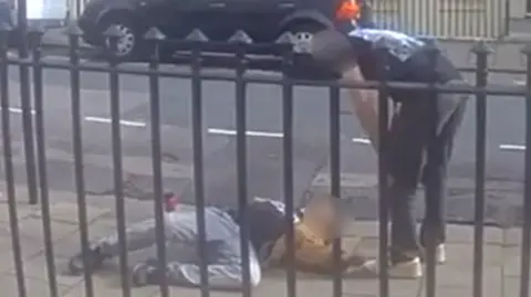 Supplied A man passed out in the street. Another man is stood over him.
