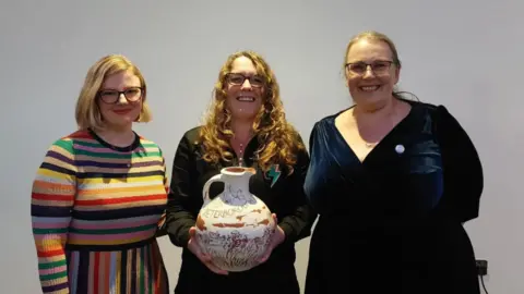 Lizzy Dening, Lauren Kendrick and Hilary Steele standing in a row smiling into the camera. They are standing in front of a white wall. Lauren Kendrick, who is in the middle and has long, red hair, glasses and is wearing a brown top, is holding a large ceramic jug - her award for being named last year's Peterborough Poet Laureate.