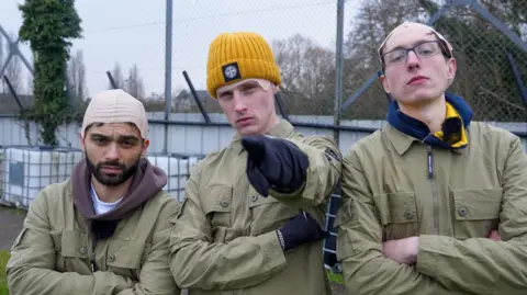 Alfie Indra, Ellis Platten and a third man all standing next to each other at a small football ground. They are all wearing green jackets and bald caps. They have serious expressions on their faces.