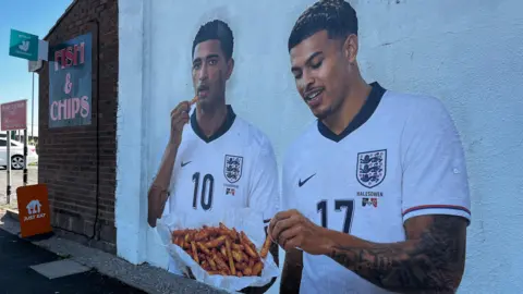 A mural on the side of a white building shows England footballers Jude Bellingham and Morgan Rogers eating from a bowl of orange chips.