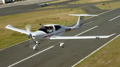 A white plane with blue and red stripes flies above a tarmac runway on a sunny day.