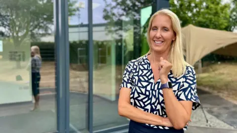 Hazel Shearing / BBC Vicky Orsmond is seen standing outside a modern glass-fronted building, wearing a patterned short-sleeved shirt and a smartwatch. A reflection is visible in the window behind, with trees, a canopy structure and landscaped grounds in the background.
