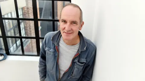 A balding man, wearing a grey t-shirt and blue denim jacket, leans casually against a whitewashed wall, beside a window with black frames.