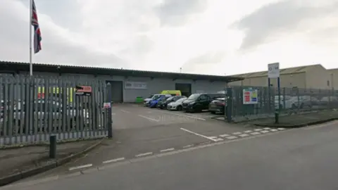 Google Maps The picture has been taken from a road, and shows a grey corrugated iron building which is as long as the photo. There are grey fences in the foreground, with cars parked behind it. 