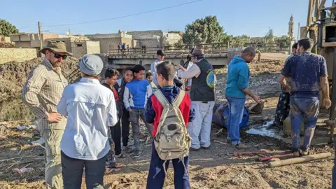 Suzanne Onstine Ερευνητές στέκονται ανάμεσα σε παιδιά σε έναν αρχαιολογικό χώρο στην Αίγυπτο