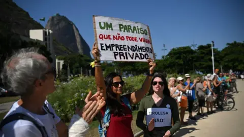 CARL DE SOUZA/AFP via Getty Images A number of people of different ages, some of them holding placards and signs, clap during a protest held at the foot of Sugarloaf Mountain in Rio de Janeiro in March 2023. One sign reads in Portuguese "No to the zipwire" while another reads "A paradise which can't become a private enterprise".