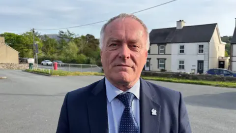 Councilor Edgar Wyn Owen looks at the camera, he is wearing a navy suit, light blue shirt and navy tie. He has grey short hair. Behind him houses are visible.