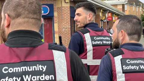 A team of three men in maroon stab vests with radios attached patrol, walking away from the camera as the lead man with black hair turns his head to look back.