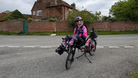 Tom Marriage Chloe can be seen sat at the front of her dual purpose bicycle.
The bike is in the middle of a road.
She is wearing a pink and black outfit.
In the background is a wall and house, with general foliage dotted around.
