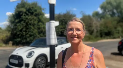 Owen Sennitt/BBC A woman smiles towards the camera as a Mini Cooper car passes behind her on a sunny day.