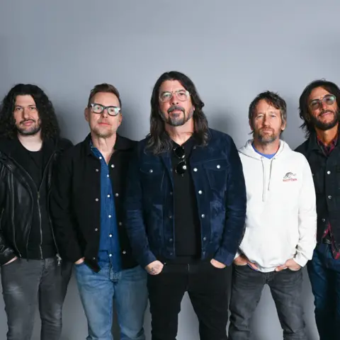 BBC Five members of the Foo Fighters band stand side by side looking directly at the camera, hands in pockets. 