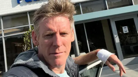 A man with short grey hair is resting his bandaged hand while standing in front of a hospital building.