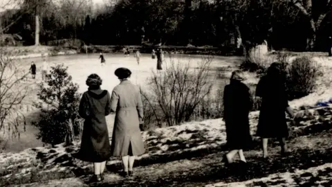 Judith Hodsdon Four women, standing with their backs to the camera, by a lake, that is frozen over, with people skating on it. They are all wearing coats. The path has snow on it, and there are trees all around the lake. The image is black and white.