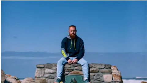 Jarred Karp A man sat on a rock staring at the camera. He has ginger hair and a beard. He is wearing a blue hoodie and blue jeans. 