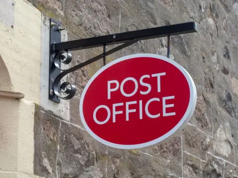 Getty Images Oval sign hanging from a stone wall with the words "Post office" in white writing on a red background.