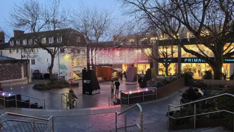 BBC A view of Hay Hill, looking towards Haymarket, showing a paved, stepped area with chrome handrails and seating areas, and a pedestrianised street beyond with a turquoise, illuminated Primark sign across a shop front.